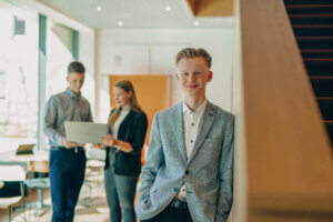 Collin angelehnt an Wand, im Hintergrund zwei Azubis stehend mit Laptop in der Hand