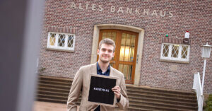 Azubi steht vor Bankgebäude. In seiner Hand ein Schild mit der Aufschrift "Marktfolge".