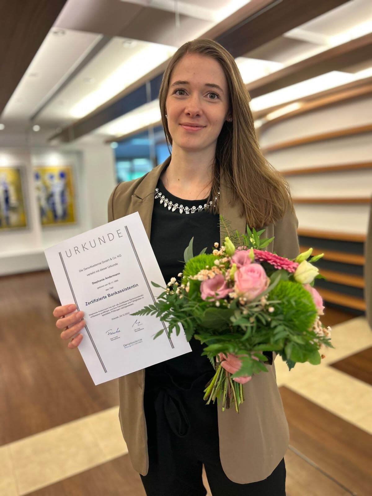 Stephanie nach bestandener Prüfung mit Blick in die Kamera, eine Urkunde in der einen und einen Blumenstrauß in der anderen Hand